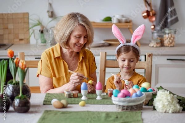 Fototapeta Happy easter family elderly grandmother and little granddaughter with rabbit ears are preparing for the holiday to paint eggs