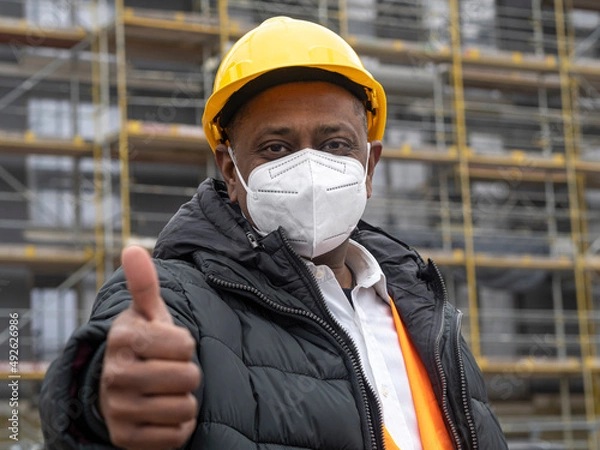 Obraz Successful Indian construction worker wearing a protective ffp2 mask posing on construction site showing thumbs up gesture