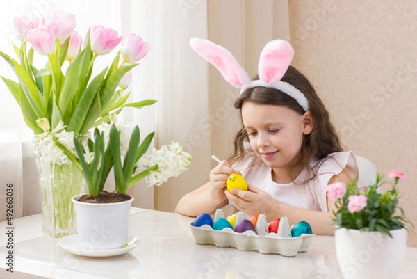 Fototapeta Cute little girl wearing bunny ears and drawing funny faces on Easter eggs.