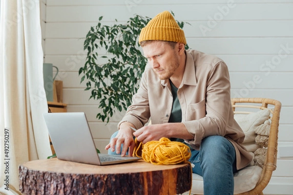 Fototapeta Young serious man looking at laptop. Man learning new hobby, knitting on needles. Knitting project in progress.