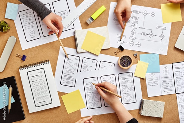 Obraz Three web UX designers working together at a busy desk with numerous mobile app wireframe sketches and user flow. Top view.