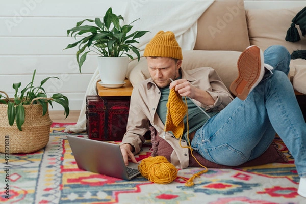 Fototapeta Young serious man looking at laptop. Man learning new hobby, knitting on needles. Knitting project in progress.