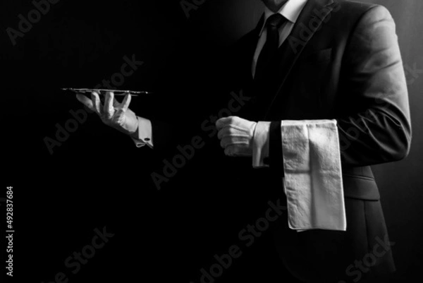 Fototapeta Portrait of Butler or Waiter in Dark Formal Suit and White Gloves with Napkin Over Arm Holding Silver Serving Tray.