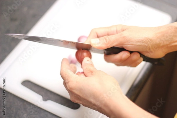 Fototapeta A woman's hand is slicing shallots on a white plastic cutting board.