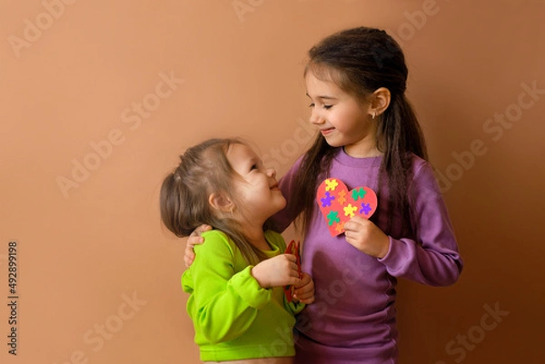 Obraz Girls look at each other, holding handmade cards with puzzles in their hand. The eldest child teaches his sister support, care and love by his own example. Symbol of autism day. No discrimination
