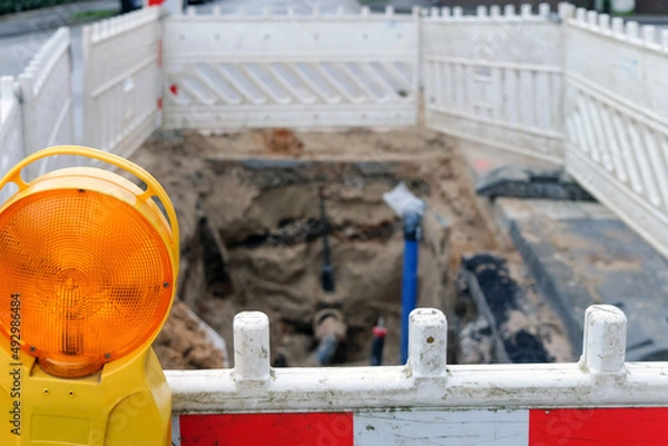Fototapeta Offene Baugrube mit Wasseranschluss und anderen Versorgungsleitungen im unscharfen Hintergrund in einer Straße, mit Absperrbarken, Schutzgitter und Baustellenlampe im Vordergrund, selektiver Fokus