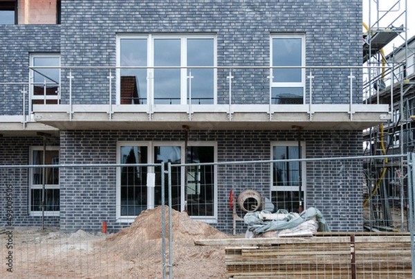 Fototapeta Frontansicht: Detail einer Baustelle eines neu gebauten Mehrfamilienhauses hinter einem Bauzaun, mit Zementmischer, Material, Brettern, Sand, Kies, Werkzeug, Gerüst, selektiver Fokus, Copy Space