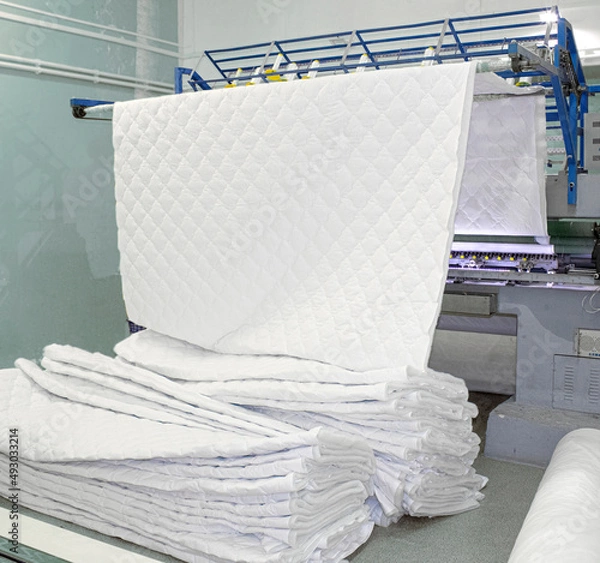Fototapeta huge sewing machine in the workshop produces a sheet of quilt, mattress pad, pillow covers, bedspreads. Multi-needle machine embroiders a pattern with threads on a white cotton fabric and winterizer