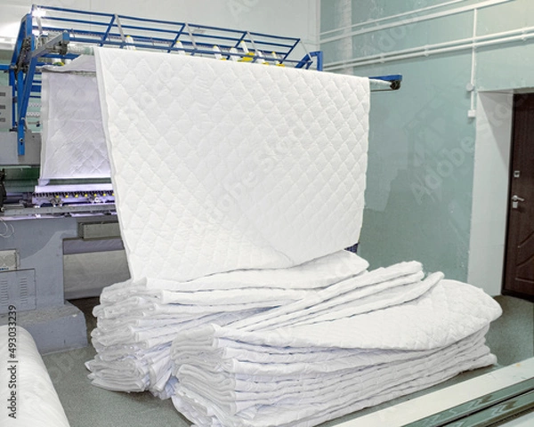 Obraz huge sewing machine in the workshop produces a sheet of quilt, mattress pad, pillow covers, bedspreads. Multi-needle machine embroiders a pattern with threads on a white cotton fabric and winterizer