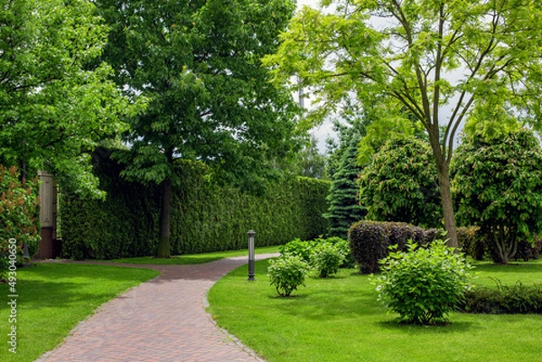 Fototapeta iron lantern ground garden lighting of park with crescent walkway paved with brick stone tiles in backyard among plants, bushes and trees surrounded by turf green lawn on sunny summer day, nobody.