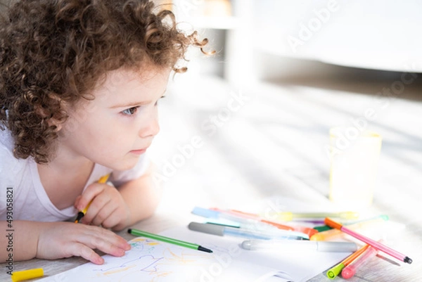 Fototapeta curly child girl drawing with colored markers lying on floor in living room at home
