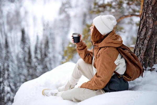 Fototapeta Calm redhead caucasian woman sit on snow on top of mountains relaxing, drinking hot tea from thermos, having rest after hike, travel, trip, adventure. inspiration, magnificent view landscape