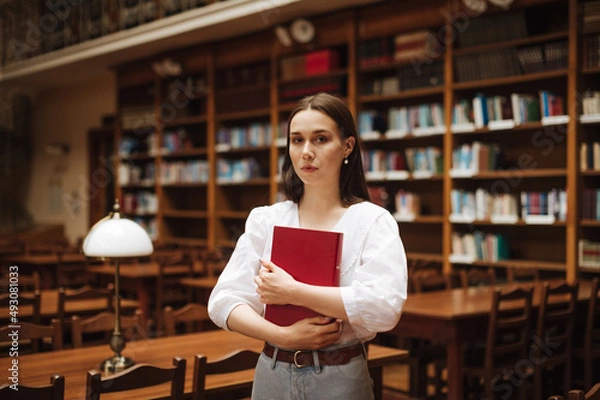Fototapeta Beautiful woman in a white blouse stands with an atmospheric cozy library with a book in her hands and poses for the camera with a serious face.
