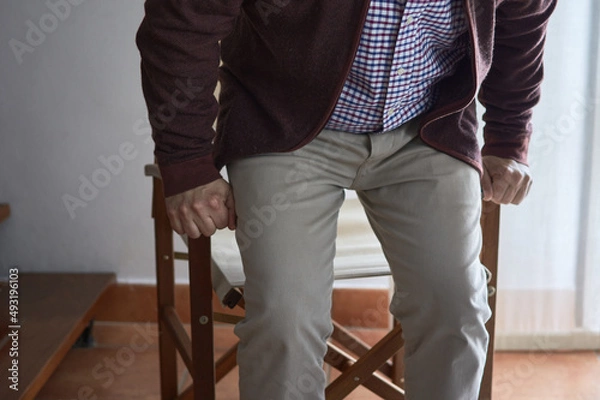 Fototapeta A mature man has difficulty getting up from an armchair in the living room of his home.