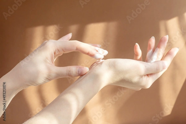 Obraz Beautiful female hands close-up apply moisturizer to delicate skin on brown background in rays of morning sun. Rubbing cosmetics, lotion into hands. Concept of care and beauty.