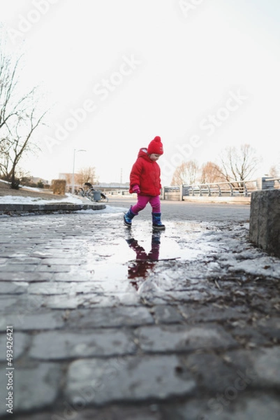 Fototapeta happy baby girl in boots and red jacket run and play in water puddle. funny cheerful toddler girl playing outdoors