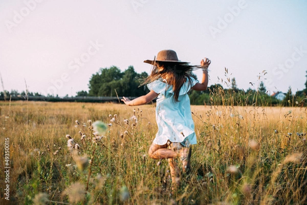 Fototapeta Kind mit langen Haaren in hellblauen Kleid und Hut rennt freudig über eine Blumenwiese im Sommer