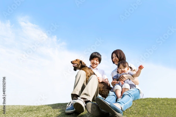 Obraz 青空を背景にペットの犬を抱き芝に座る笑顔の若い夫婦と幼い娘。家族,幸せ,愛情,育児とペットのイメージ