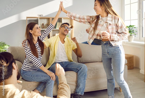 Fototapeta Happy excited young women give high fiving each other while having fun at home with friends. Positive cheerful women together with their boyfriends have fun together at casual gathering at home.
