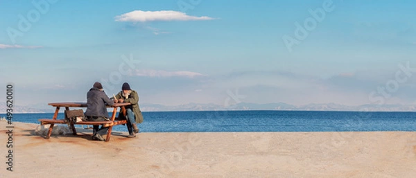 Fototapeta Old men sits on a bench by the sea. One man suggesting a book the other man. They are retired people. Living peaceful, quiet seaside town.	Empty space for text. Cop paste, web banner.
