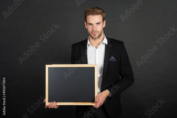 Obraz Serious guy in formal suit holding empty school blackboard copy space dark background, teacher