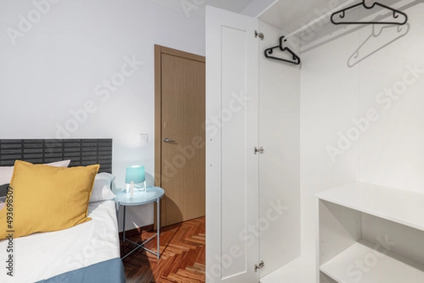 Fototapeta Open white wardrobe with shelves, coat rack and black hangers in bedroom with round blue nightstand and mahogany herringbone parquet flooring