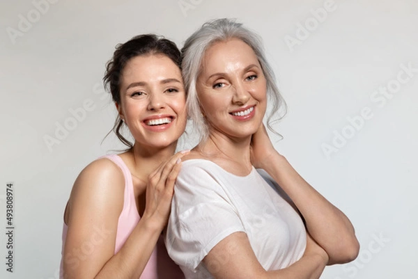 Fototapeta Young woman and smiling senior lady hugging portrait. Modest shy millennial daughter covering face with palm cuddling confident experienced aged mother from back. Trust protection, love support