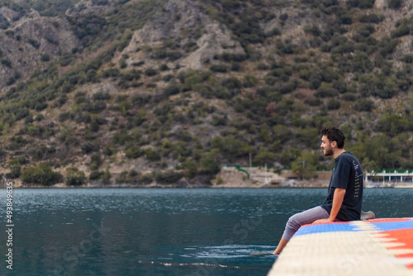 Fototapeta Barefooted young man sitting on colorful dock. He is happy. He puts his feet in sea.
