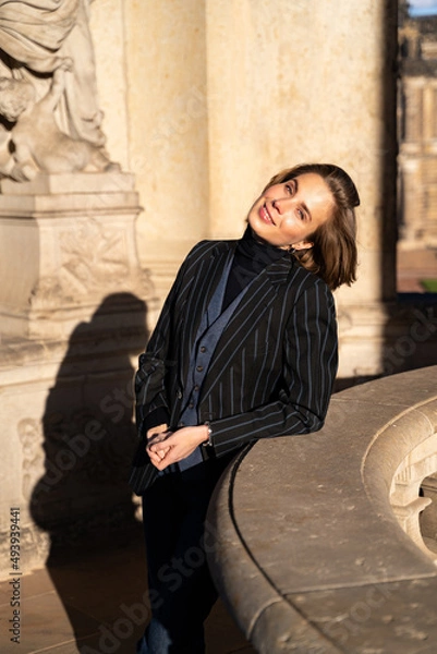 Fototapeta Young blond cheerful happy smiling business woman in retro styled striped jacket and wristwatch looking in camera leaned on historic stone building with sculpture in Dresden on sunny day. Lifestyle