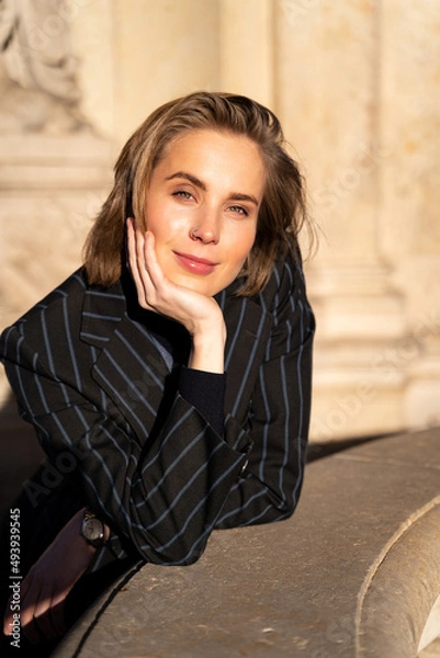 Fototapeta Portrait of young blond cheerful happy smiling woman with peirced nose, in retro styled striped classic jacket and wristwatch looking in camera leaned on hand near historic stone building on sunny day