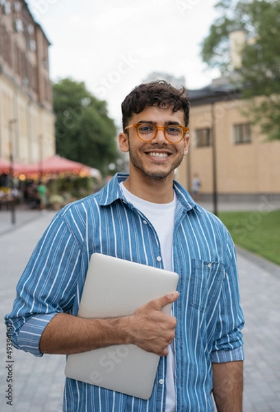 Fototapeta Authentic portrait of young handsome Indian man wearing stylish eyeglasses standing on the street. Smiling asian student holding laptop looking at camera in university campus. Education concept 