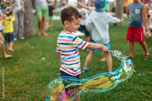 Fototapeta the child inflates large soap bubbles. Street animation program, happy child on a green lawn. Summer vacation. A boy in bright striped clothes with soap bubbles. Large portrait, horizontal.