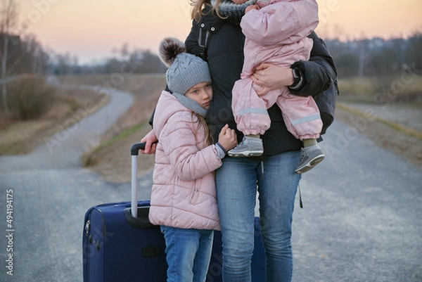 Fototapeta A refugee woman walks along the road with a suitcase on wheels and holds a child in her arms.