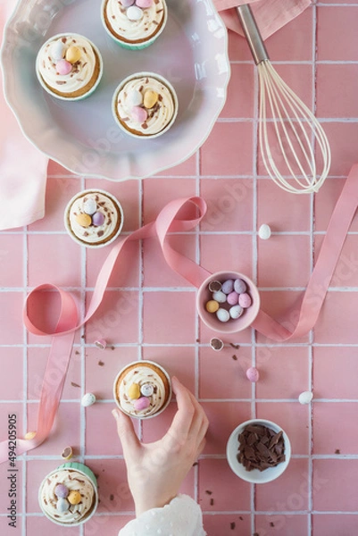 Fototapeta Flat lay Easter baking concept image. Easter theme cupcakes image with chocolate and baking equipment included in frame. Woman's hand reaches into frame to take a frosted cup cake. Pastel pink colours