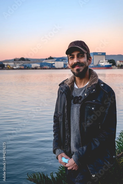 Fototapeta Handsome and happy young man with beard, snapback and jacket standing with sunset over the lake, shipyard and harbor in the back, Valdivia, Chile