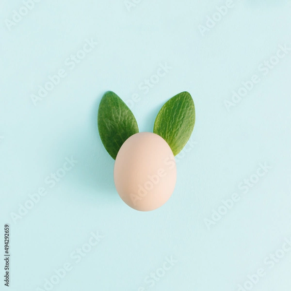 Obraz Easter bunny festive idea with an egg and fresh green leaves. Holiday spring season concept, minimal flay lay composition on pastel blue background.
