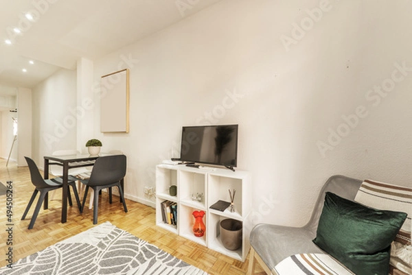 Fototapeta Corner of a living room with an armchair next to a bookcase with flat screen TV and a square glass and iron dining table