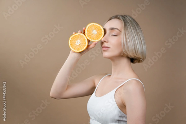 Fototapeta Slim woman with beautiful face complexion holding oranges halves in hands. Model in white lingerie posing on beige background, advertising healthy lifestyle and fitness concept.