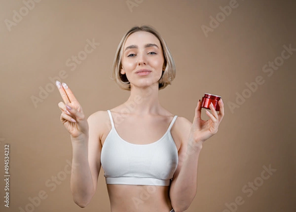 Fototapeta Young woman with healthy face complexion holding small red container with skincare cream. Model in white sport top posing on beige background, advertising cosmetic products.