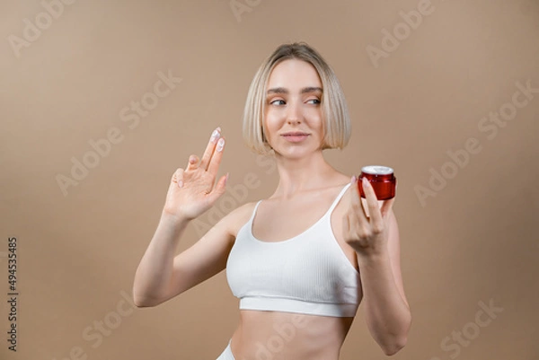 Fototapeta Young woman with healthy face complexion holding small red container with skincare cream. Model in white sport top posing on beige background, advertising cosmetic products.