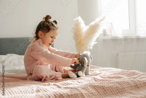 Fototapeta A cheerful little girl on the bed after waking up plays with her toy elephant