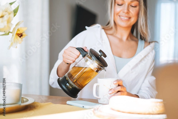 Fototapeta Young beautiful happy woman with blonde long hair in white shirt drinking herbal tea on dinner table in bright interior at the home