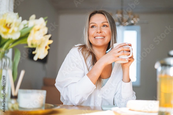 Fototapeta Young beautiful happy woman with blonde long hair in white shirt drinking tea with bouquet of yellow flowers in vase on dinner table in bright interior at home
