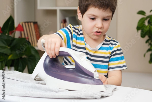 Fototapeta A boy of 4 years learns to hold an iron, iron clothes on an ironing board. Helper, help children to care for the house.