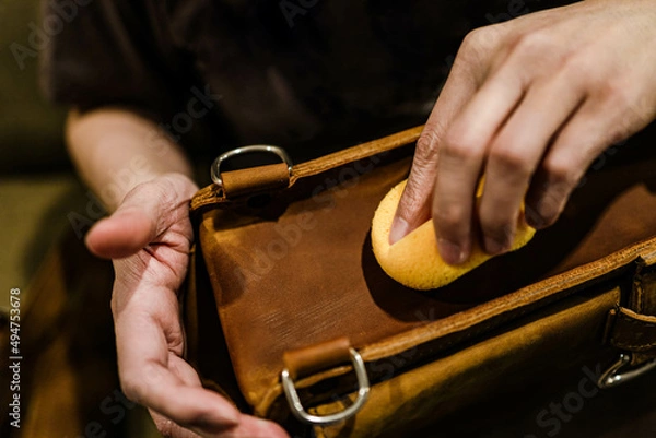Fototapeta Professional leather care service concept. Closeup hand with yellow soft sponge cleaning surface of the vintage brown bag, depth of field. Polish waxing protection from dust and dirt.