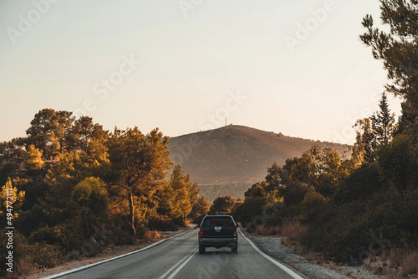Fototapeta A pick up truck driving on a n asphalt road going through rocks and trees in the countryside of Rhodes, Greece