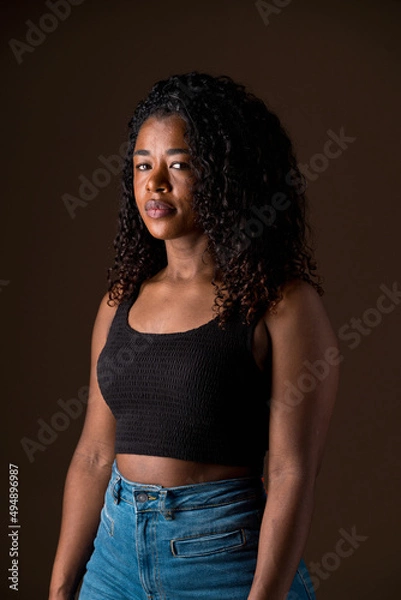 Obraz Mid waist portrait of african american woman with dark hair and casual clothes. Vertical side view of african woman with quiet expression in brown background. People and color concept.