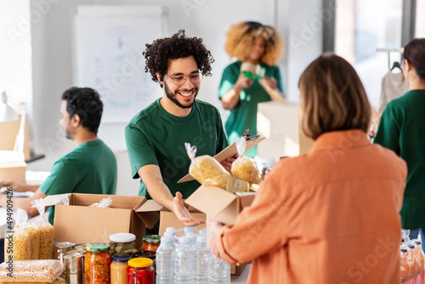 Fototapeta charity, donation and volunteering concept - happy smiling male volunteer with clipboard and woman taking box of food at distribution or refugee assistance center