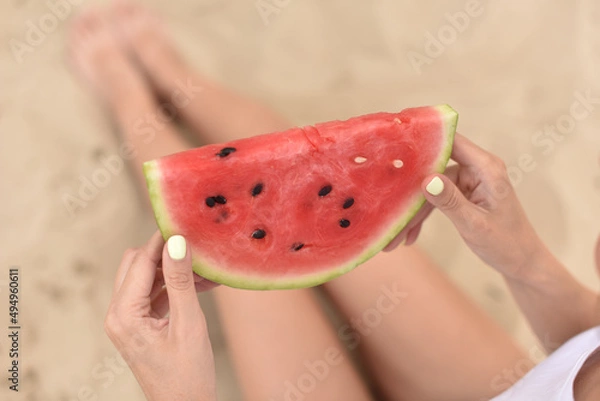 Fototapeta girl holds red watermelon slice in hands. picnic on the beach. young woman sit on sand in summer vacation. shallow depth of field