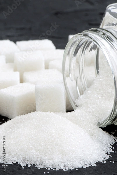 Fototapeta Glass jar with sugar black background.Diabetes. Scattered white sugar on a black table. Copy space. Place for text.Extra calories. Vertical orientation